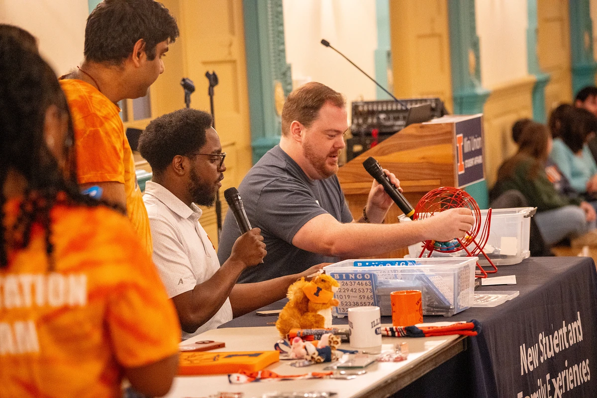 Three people are speaking into microphones while hosting a bingo event at a table with various items.